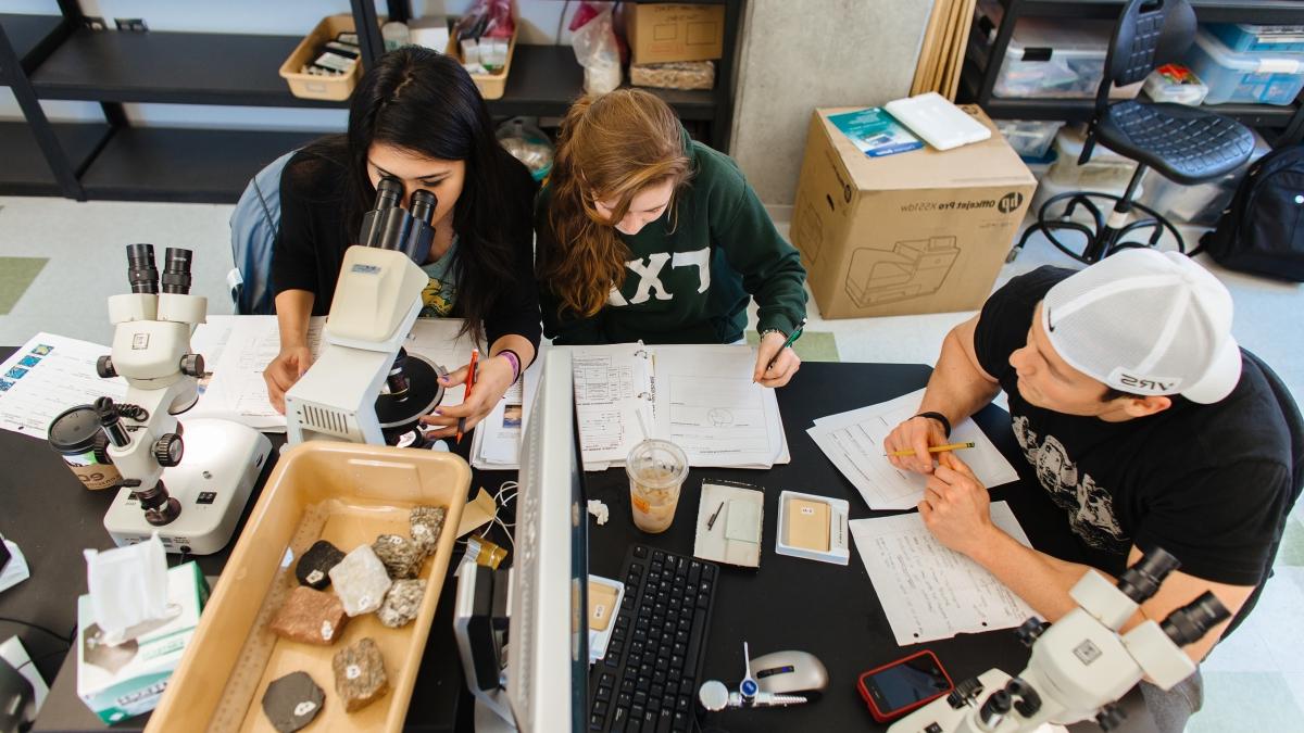 两名学生做笔记，另一名学生在显微镜下观察岩石样本